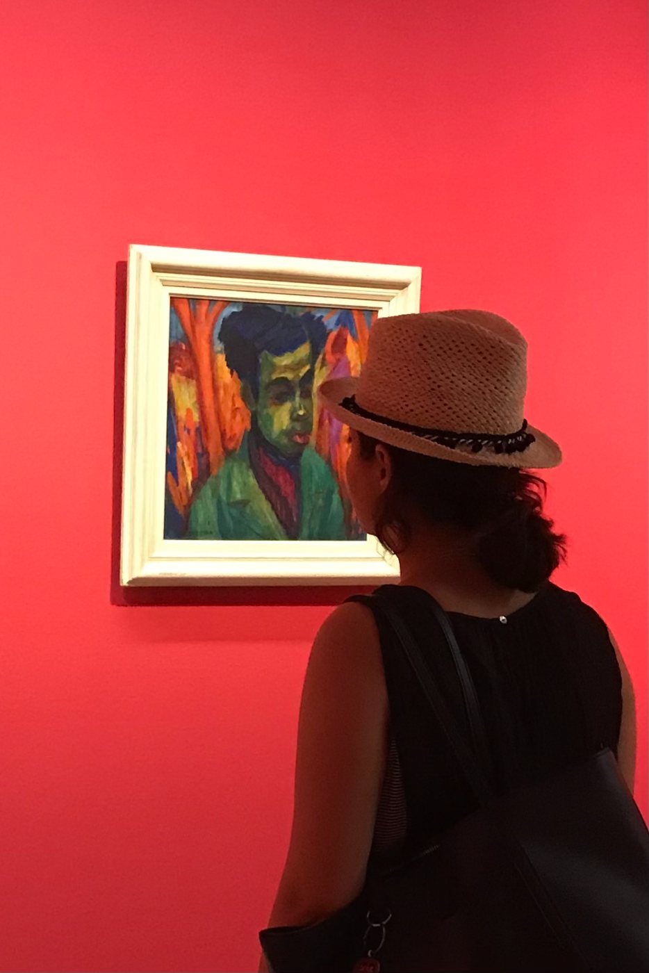 Negin in a straw hat looking at an expressionist portrait on a red gallery wall